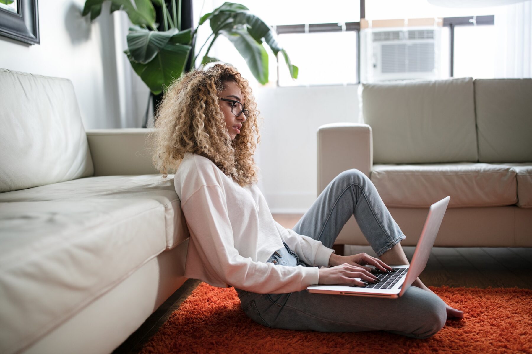 A person with curly hair and glasses sits on an orange rug, leaning against a white sofa, typing on a laptop in a bright living room with large windows and green plants.